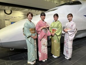 Photograph of four young women in traditional Japanese attire standing in front of a museum display of a Shinkansen high-speed train.
