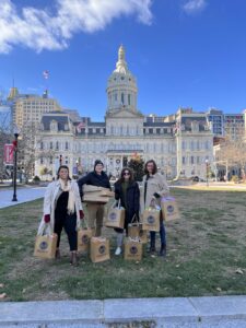 Photograph of four members of the Northeast Maglev marketing team standing in front of Baltimore City Hall and holding bags of toys for the Mayor's annual Toys for Tots charity drive.